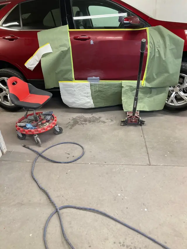 Red SUV door masked for paint in the bay, with a creeper and jack beside it